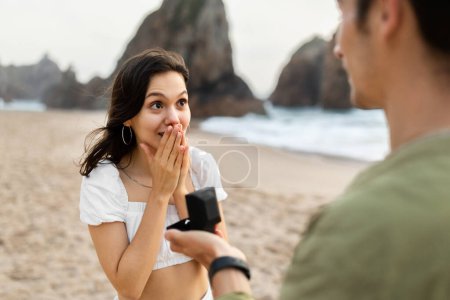 Téléchargez les photos : Homme romantique faire une demande en mariage à sa belle femme surprise avec bague dans une boîte cadeau tout en marchant sur la plage et avoir rendez-vous sur le littoral. Épouse-moi concept - en image libre de droit