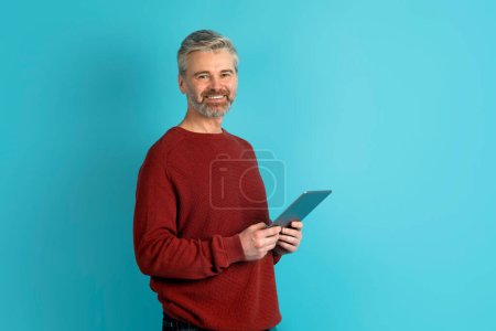 Téléchargez les photos : Joyeux beau barbu aux cheveux gris homme d'âge moyen en pull rouge en utilisant une tablette numérique et souriant, isolé sur fond bleu, espace de copie. Chatbot, assistant en ligne, IA dans la vie quotidienne - en image libre de droit