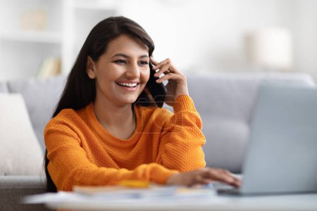 Photo for Closeup of cheerful pretty long-haired millennial indian woman wearing orange sweater entrepreneur using laptop and talking on cell phone at home, working online, copy space. Remote job - Royalty Free Image