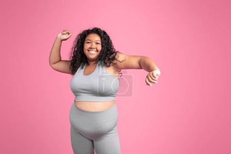 Téléchargez les photos : Femme oversize afro-américaine excitée dans la danse de vêtements de sport, célébrant la formation réussie, acclamant et levant les mains vers le haut, fond de studio rose, bannière, espace de copie - en image libre de droit