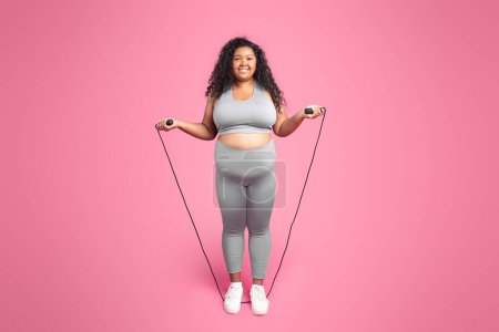 Foto de Mujer positiva cuerpo negro positivo en ropa deportiva posando con saltar la cuerda, haciendo entrenamiento de fitness, de pie sobre fondo rosa, longitud completa, espacio de copia - Imagen libre de derechos