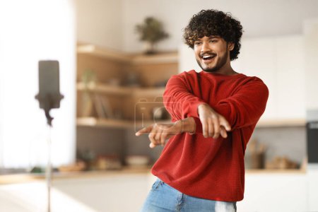 Photo for Handsome Indian Male Blogger Dancing At Smartphone Camera While Recording Video At Home, Cheerful Eastern Guy Creating Content For His Social Networks, Using Mobile Phone On Tripod, Copy Space - Royalty Free Image