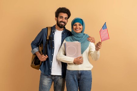 Photo for Cheerful muslim students young man and woman in hijab holding american flag over beige studio background, smiling arabic couple carrying backpacks and workbooks, studying in USA, copy space - Royalty Free Image