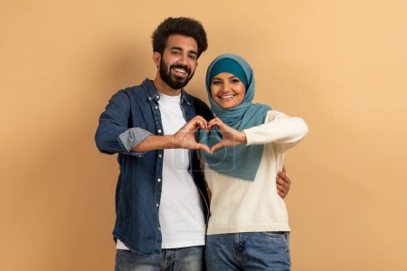 Photo for Portrait of romantic muslim couple making heart gesture with their hands, loving arab man and woman in hijab celebrating Valentines Day, looking at camera and smiling, posing over beige background - Royalty Free Image