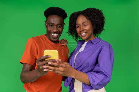 Photo for Happy young african american guy showing smartphone to woman, recommending app, isolated on green studio background. Relationships, social networks, blog and device, watch funny video - Royalty Free Image