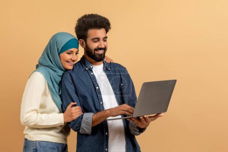 Téléchargez les photos : Super site web. Couple musulman joyeux en utilisant l'ordinateur portable ensemble, sourire homme arabe et femme dans Hijab Shopping en ligne ou en naviguant sur Internet tout en se tenant debout sur Beige Studio fond, Copier l'espace - en image libre de droit