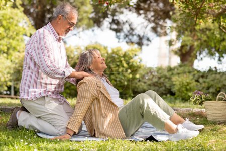 Téléchargez les photos : Heureux vieux mari européen faisant massage des épaules à la femme sur plaid sur herbe verte, couple profiter pique-nique ensemble dans le parc, en plein air. Soins, amour, rendez-vous romantique, mode de vie et week-end, temps libre - en image libre de droit