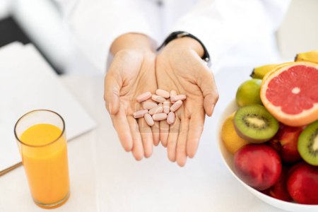 Foto de La nutricionista médica europea adulta en bata blanca en la mesa con frutas orgánicas y jugo tiene pastillas en las manos en la clínica de la oficina. Recomendación atención médica, nutrición adecuada, pérdida de peso, dieta - Imagen libre de derechos