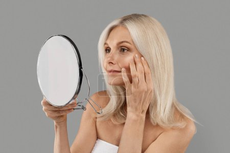 Photo for Skin Aging. Attractive Mature Woman Holding Mirror And Looking At Her Wrinkles Near Eyes, Beautiful Senior Female Checking Face, Noticing Age Changes, Standing Wrapped In Towel On Grey Background - Royalty Free Image