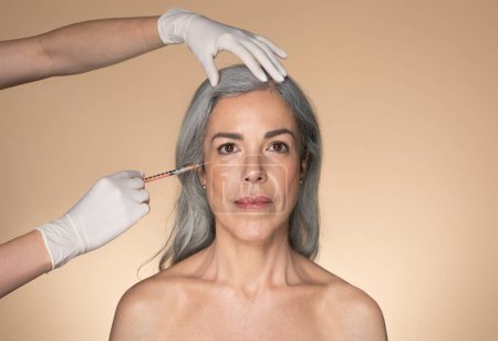 Photo for Cosmetologist doctor making botox injection for eyes area to senior grey-haired woman, beautiful aged lady getting anti-wrinke treatment over beige studio background - Royalty Free Image