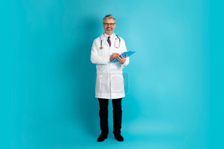 Photo for Positive cheerful friendly handsome grey-haire bearded mature doctor wearing eyeglasses taking anamnesis on blue background, holding medical chart and smiling at camera, copy space. Medical staff - Royalty Free Image