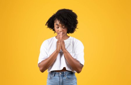 Téléchargez les photos : Sérieuse femme noire millénariste dans un geste occasionnel de prière avec les mains, isolée sur fond de studio orange. Espoir, souhait devenu réalité, demande, émotions humaines, style de vie, annonce et offre - en image libre de droit