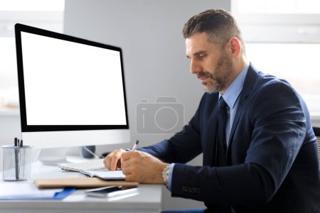 Téléchargez les photos : Homme d'affaires sérieux d'âge moyen à table avec ordinateur avec écran vide prenant des notes, planifiant démarrage, travaillant dans le bureau, maquette. Dispositif pour les entreprises et le travail, créer une idée - en image libre de droit