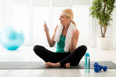 Photo for Smiling senior woman in earphones having video call on smartphone while resting after training, happy mature female talking with fitness coach, sitting on yoga mat in living room, copy space - Royalty Free Image