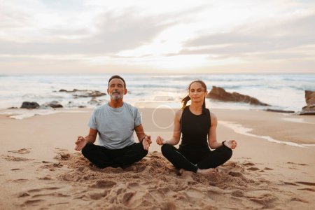 Foto de Serious senior caucásico dama y hombre en ropa deportiva practicar yoga y ejercicios de respiración con los ojos cerrados en la playa por la mañana, al aire libre. Meditación juntos, descanso, relax y paz - Imagen libre de derechos