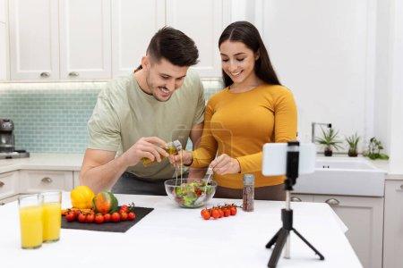 Photo for Beautiful young family husband and wife cooking together at home. Handsome man and pretty woman standing by kitchen desk, making healthy salad, checking nice recipe online, using smartphone - Royalty Free Image