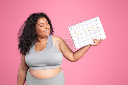 Photo for Sports and menstruation. Smiling black overweight woman in sportswear holding calendar, posing on pink studio background. Weight loss and body care time management, health care - Royalty Free Image