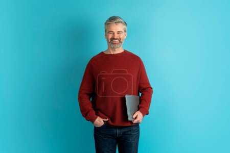 Photo for Entrepreneurship and technologies. Cheerful handsome middle aged man startup founder or small business owner managing business operations via his laptop, blue studio background, copy space - Royalty Free Image