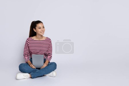 Photo for Cheerful pretty brunette hispanic young woman in casual outfit sitting on floor over grey studio background, using digital tablet, looking at copy space. Online assistant, chatbot in daily life - Royalty Free Image