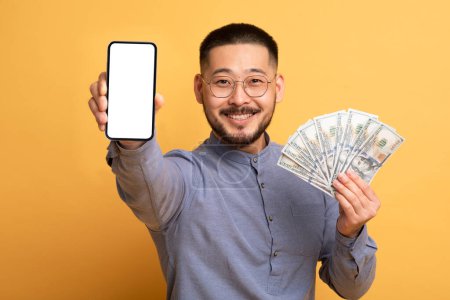 Photo for Mobile Banking. Excited Asian Guy Showing Blank Smartphone And Holding Money Cash, Handsome Millennial Man Recommending New E-Wallet Application, Standing On Yellow Studio Background, Mockup - Royalty Free Image