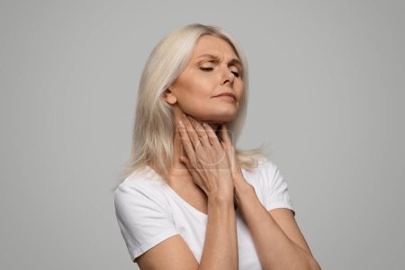 Portrait of unhappy sad sick mature woman suffering from sore throat, ill senior lady touching neck with hands, feeling ache, standing isolated over grey studio background, copy space