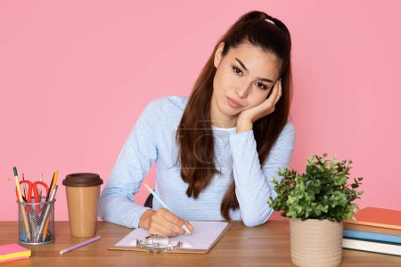Photo for Bored attractive latin brunette young woman in smart casual outfit secretary sitting at office table, holding pen, taking notes, leaning on her arm, feeling tired, overworked, pink background - Royalty Free Image