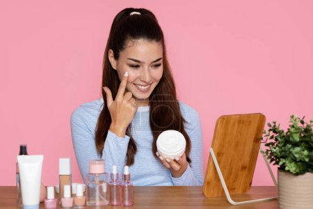 Photo for Attractive brunette long-haired latin young woman in casual outfit looking at mirror, holding facial cream, applying sunscreen and smiling, pink background, copy space. Sun protection for face - Royalty Free Image