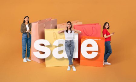 Photo for Sales Season Offer. Three Happy Latin Women With Phone Shopping Online In App Near Big Paper Bags Over Orange Studio Background. Collage With Buyers Showing Like And Shoppers Advertising Discount - Royalty Free Image