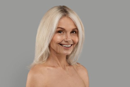 Téléchargez les photos : Portrait de sourire attrayant femme mûre avec les épaules nues posant sur fond de studio gris, heureuse belle femme âgée avec maquillage naturel et cheveux blonds regardant l'appareil photo, espace de copie - en image libre de droit