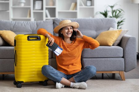 Photo for Happy cheerful attractive young eastern woman wearing casual outfit and wicker hat tourist sitting on floor with passport, tickets, luggage talking on phone at home, calling taxi, copy space - Royalty Free Image