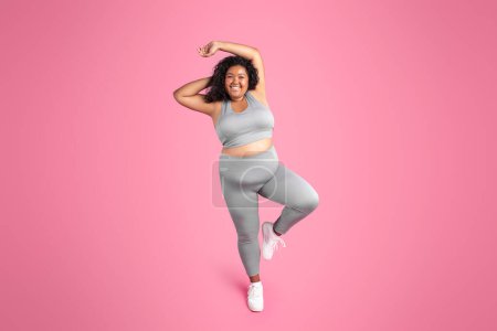 Foto de Mujer gordita afroamericana positiva en ropa deportiva descansando después del entrenamiento y disfrutando del resultado del entrenamiento, fondo de estudio rosa, longitud completa. Fitness, pérdida de peso y cuidado corporal - Imagen libre de derechos