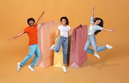 Photo for Great Sales. Joyful Diverse Young Man And Women Near Big Bags Celebrating Black Friday Shopping Offer, Jumping On Orange Background, Collage. Full Length Shot Of Emotional Shopaholics Trio - Royalty Free Image