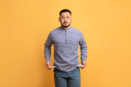 Photo for Poverty Concept. Portrait Of Upset Asian Guy Showing His Empty Pockets And Looking At Camera, Broke Poor Millennial Man Standing Over Yellow Studio Background, Suffering Financial Crisis, Copy Space - Royalty Free Image