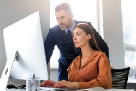 Photo for Focused employees young woman and middle aged man looking at computer screen, working on project together, sitting at desk in office interior. Teamwork concept - Royalty Free Image