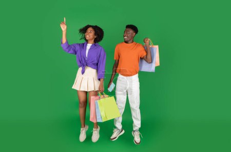 Téléchargez les photos : Souriant jeune afro-américain homme et femme boutiquiers avec des sacs d'achats saute, congeler dans l'air, pointez du doigt l'espace vide, isolé sur fond de studio vert. Temps libre, vente, shopping - en image libre de droit