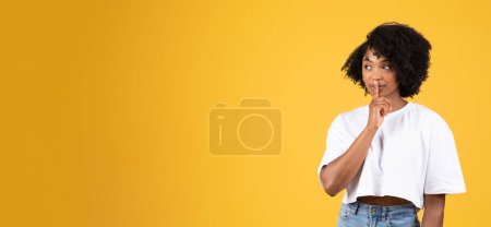 Téléchargez les photos : Positif sérieux jeune femme noire dans occasionnel faire shh geste avec le doigt, isolé sur fond de studio orange, panorama. Secret, surprise, commérages et bonnes nouvelles, émotions humaines, annonce et offre - en image libre de droit