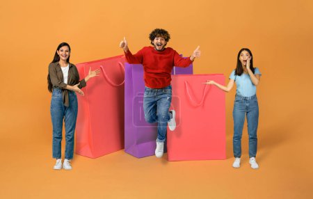 Photo for Great Sales Offer. Three Excited Indian Women And Man Posing Showing Huge Paper Shoppers, Jumping And Having Fun Advertising Discounts On Black Friday Over Orange Studio Background, Collage - Royalty Free Image