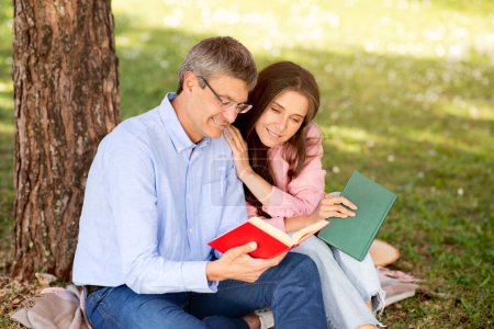 Téléchargez les photos : Beau couple d'âge mûr se reposant au parc d'été, lire le livre ensemble, heureux conjoints d'âge moyen discuter roman intéressant tout en se relaxant sur la pelouse sous l'arbre, profiter des loisirs en plein air, espace libre - en image libre de droit