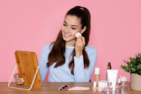 Téléchargez les photos : Joyeux sourire joyeux jolie brune jeune femme assise au bureau sur fond de studio rose, regardant miroir, blogueur beauté appliquant fond de teint de maquillage ou de fard à joues, en utilisant une éponge, espace de copie - en image libre de droit