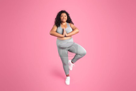 Téléchargez les photos : Équilibre de vie. Femme potelée noire calme pratiquant le yoga, faisant la pose d'arbre, s'entraînant sur fond de studio rose, espace libre. Joyeux oversize dame debout sur une jambe avec les mains serrées - en image libre de droit