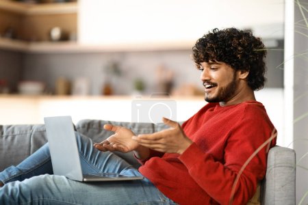 Photo for Smiling Young Indian Man Making Video Call On Laptop At Home, Handsome Millennial Eastern Male Using Computer For Web Conference While Sitting On Couch In Living Room, Enjoying Online Communication - Royalty Free Image