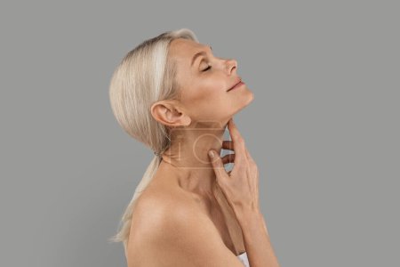 Photo for Neck Rejuvenation. Beautiful Mature Woman Touching Her Soft Skin And Smiling, Side View Shot Of Attractive Senior Female Enjoying Beauty Treatment Result, Standing Over Grey Studio Background - Royalty Free Image