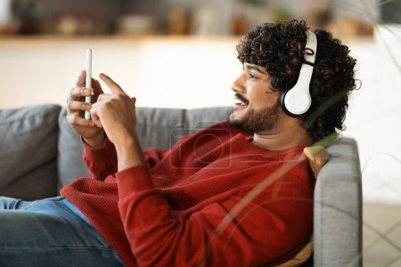 Photo for Relaxed indian guy listening music on smartphone with wireless headphones, relaxing on couch at home, enjoying favorite songs on mobile phone, downoading playlist, resting on sofa in living room - Royalty Free Image