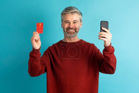 Téléchargez les photos : Joyeux beau homme d'âge moyen aux cheveux gris en tenue décontractée montrant téléphone et carte bancaire en plastique rouge, isolé sur fond de studio bleu. Services bancaires en ligne, achats, concept de commerce électronique - en image libre de droit