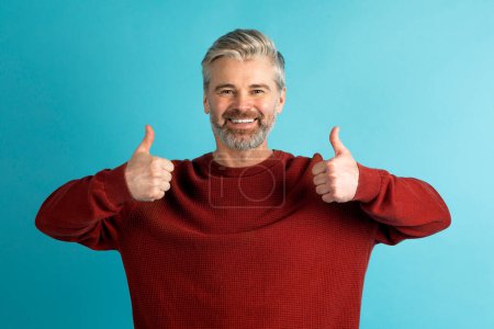 Photo for Happy positive handsome grey-haired bearded middle aged man in red sweater showing thumb ups gesture and smiling at camera on blue studio background. Human emotions and gestures concept - Royalty Free Image