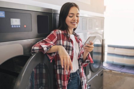 Photo for Happy Young Lady Using Cellphone While Doing Laundry, Standing Leaning On Industrial Washing Machine, Texting And Booking Laundromat Online Via Mobile Application Indoor. Laundrette App - Royalty Free Image