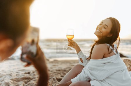Foto de Mujer tomando una foto instantánea de su novia feliz en la playa, haciendo picnic y bebiendo vino al atardecer en la costa. Señoras teniendo despedida de soltera al aire libre - Imagen libre de derechos