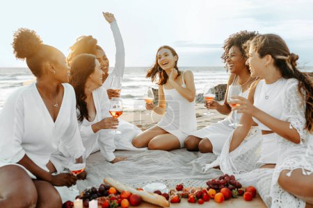 Foto de Emocionadas amigas haciendo fiesta de despedida de soltera, bebiendo vino, comiendo frutas frescas y charlando, haciendo un picnic a la orilla del mar al atardecer - Imagen libre de derechos
