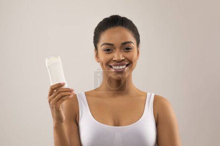 Foto de Positiva hermosa joven afroamericana dama en la parte superior blanca mostrando desodorante y sonriente, recomendando desodorante libre de aluminio para la higiene de las axilas, aislado sobre fondo gris - Imagen libre de derechos