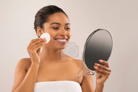 Foto de Rutina de cuidado. Feliz atractiva africana americana millennial mujer mirando pequeño espejo, utilizando almohadilla de algodón, cara de limpieza con agua micelar, utilizando tóner, aislado en gris, espacio de copia - Imagen libre de derechos